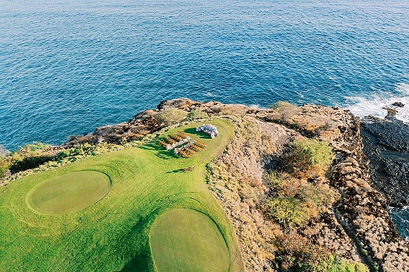 Ceremony setup with wood chairs in a semicircle around a floral arch, aisle marker florals on a cliffside lawn above ocean rocks.