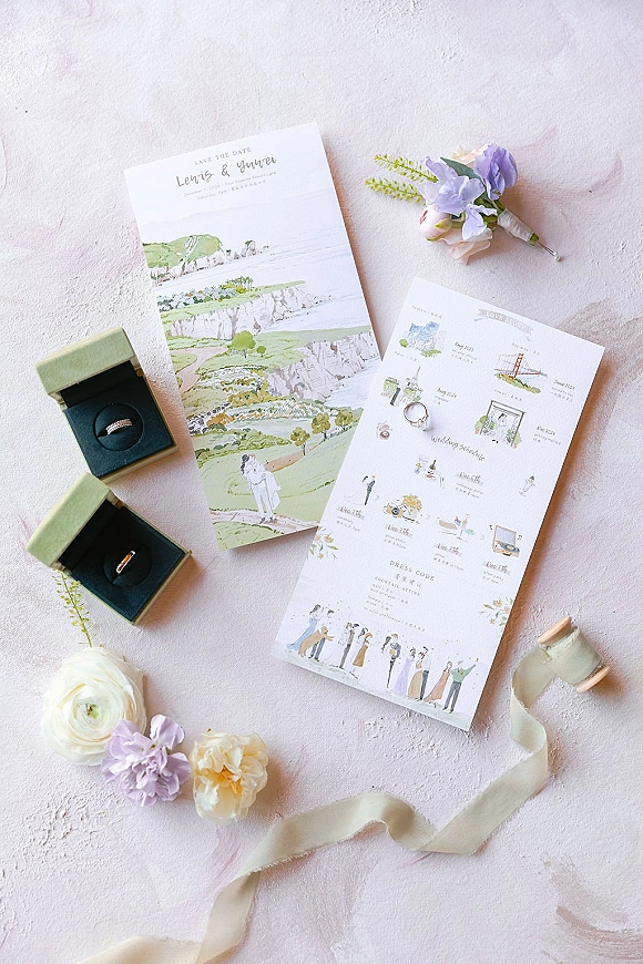 Wedding invitation suite with illustrated wedding invitations, rings in velvet boxes, boutonniere, flowers, and silk ribbon on a textured blush surface