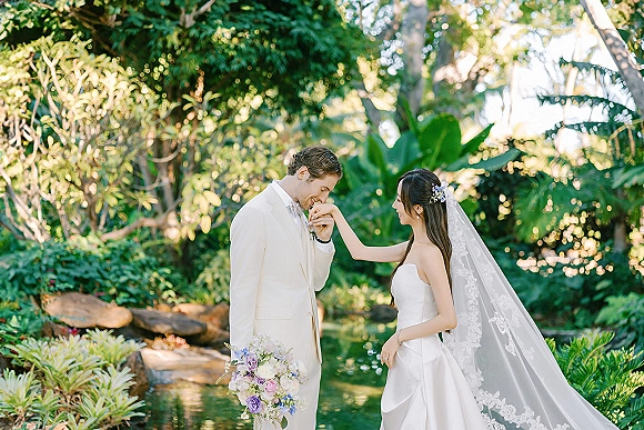 Couple portrait of groom kissing the bride’s hand beside a garden pond, her long veil and bouquet framed by tropical greenery and rocks