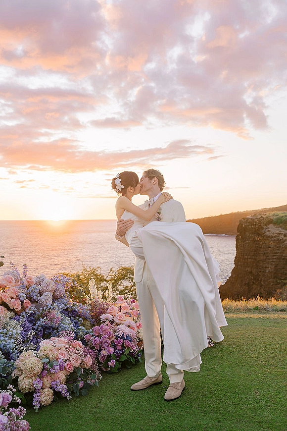 Wedding kiss portrait of groom carrying bride in a bridal lift pose, kissing at sunset by ocean cliffs with floral hair accents