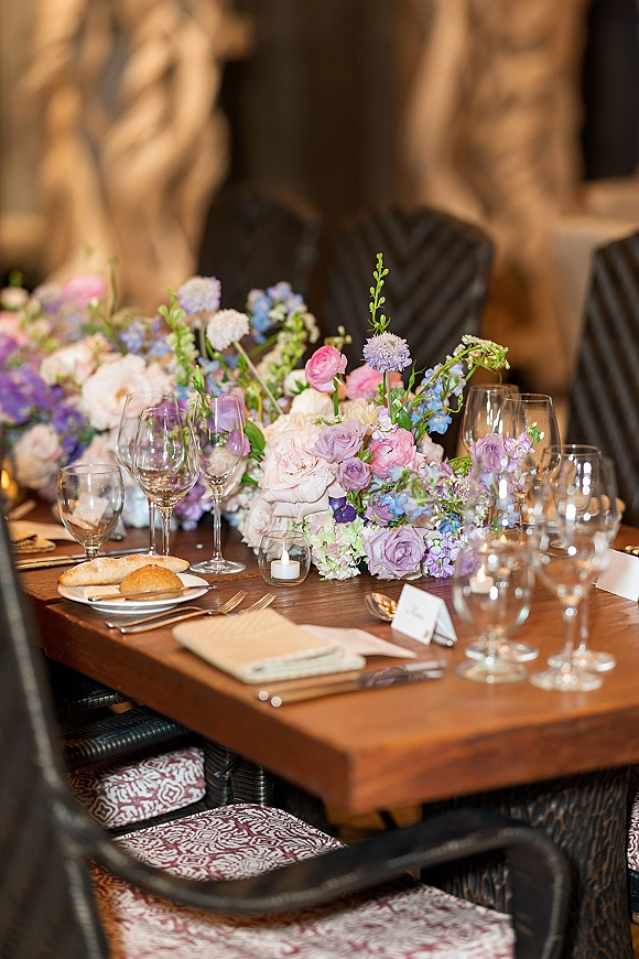 Reception tablescape with wedding table centerpiece of pastel roses and hydrangea, votive candles, wine glasses, and place cards in warm indoor light