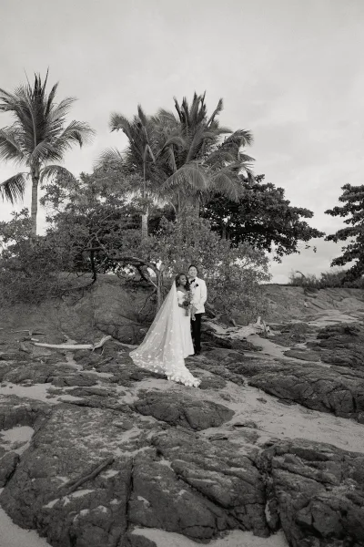 Couple portrait of bride and groom embracing on a rocky beach, her long veil trailing in the breeze with bouquet and palm trees behind
