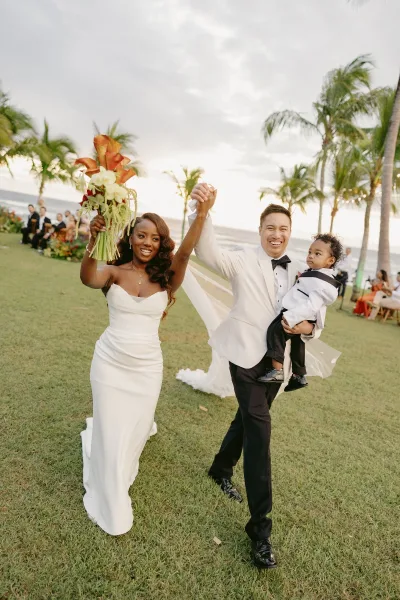 Wedding recessional as bride and groom hold hands, bride lifting bouquet and groom in white jacket, walking past guests on oceanfront lawn with palm trees
