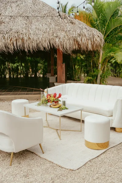 Wedding lounge area with reception lounge seating, white sofa and ottomans around a coffee table under string lights in a thatched pavilion