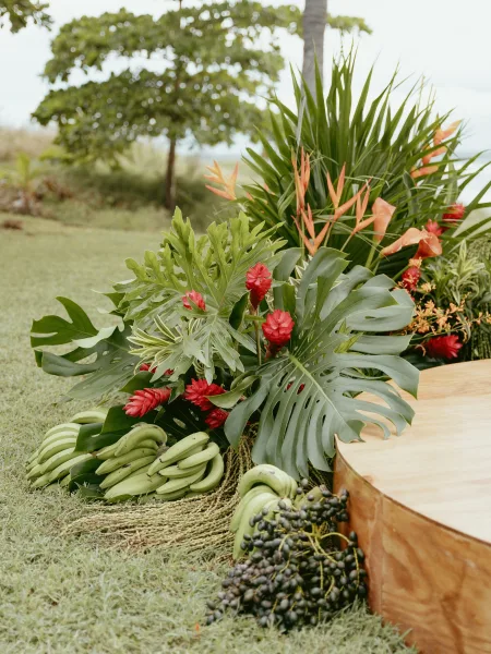 Tropical wedding florals with a monstera wedding arrangement, orchids, heliconia, and red ginger on a wooden platform on a grassy lawn
