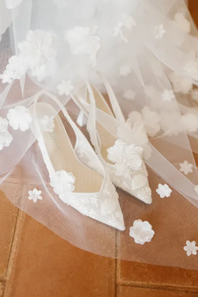 Bridal shoes in a wedding heels flat lay, white pointed toe pumps with lace and 3D florals beneath a tulle veil on terracotta tile floor