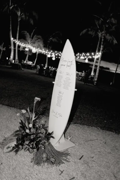 Wedding seating chart displayed on a surfboard seating chart sign with calla lilies and tropical greenery at a beachside lawn reception under string lights