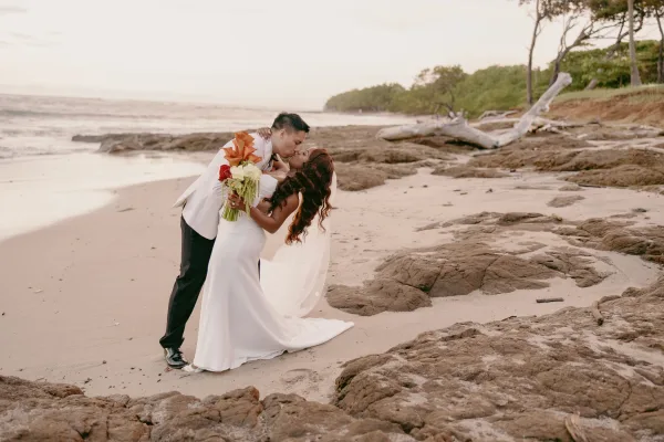 Wedding kiss portrait of groom dipping bride in veil, holding bouquet, on a sandy beach with ocean waves and rocky shoreline behind