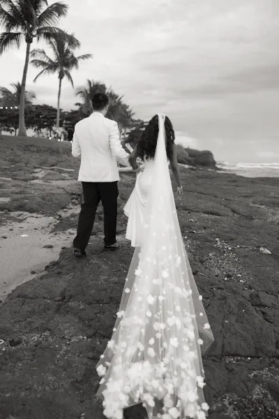 Couple portrait of bride and groom walking away holding hands on a rocky oceanfront beach, her cathedral veil trailing under palm trees