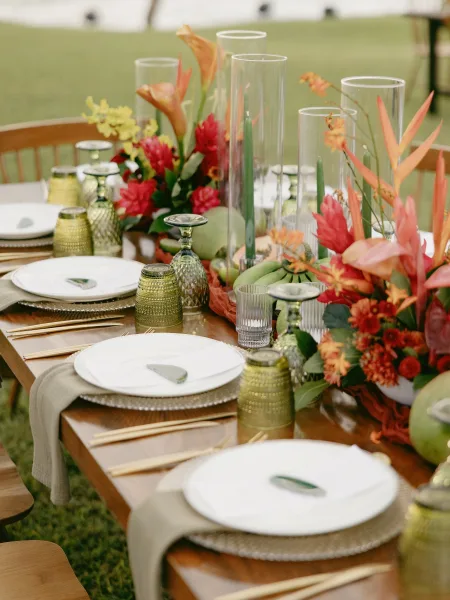 Reception tablescape with an outdoor reception table featuring tropical florals, green taper candles, gold flatware, under a tent canopy on grass lawn