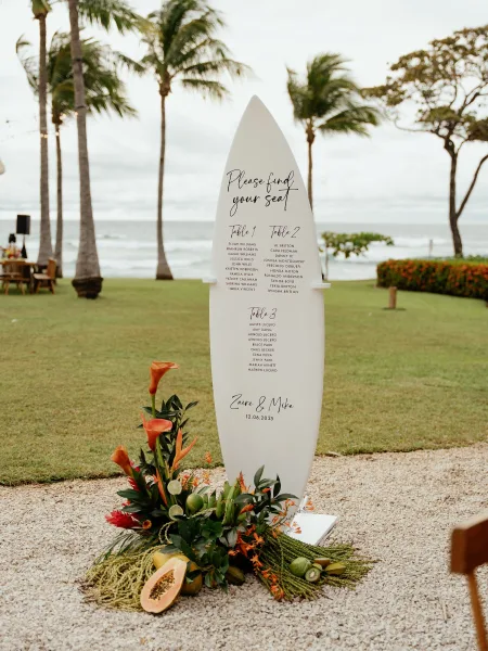 Wedding seating chart on a surfboard sign with calla lilies and tropical greenery, styled on a lawn with ocean and palm trees behind