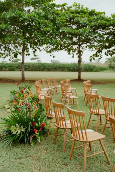 Ceremony setup with outdoor ceremony seating of wood chairs in a curved layout, aisle florals with tropical greenery on a lawn amid trees