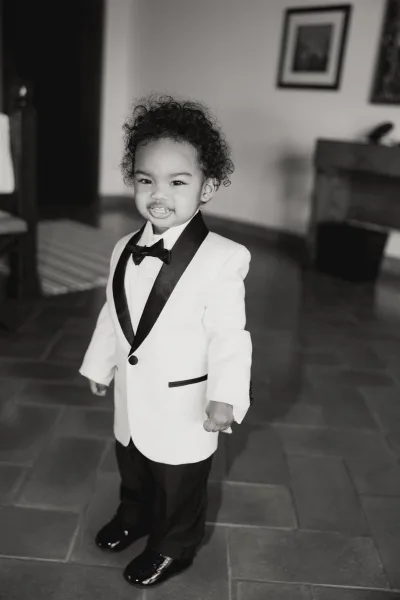Ring bearer portrait of a little boy in a white tuxedo jacket with black lapels and bow tie, standing in an indoor room with framed art