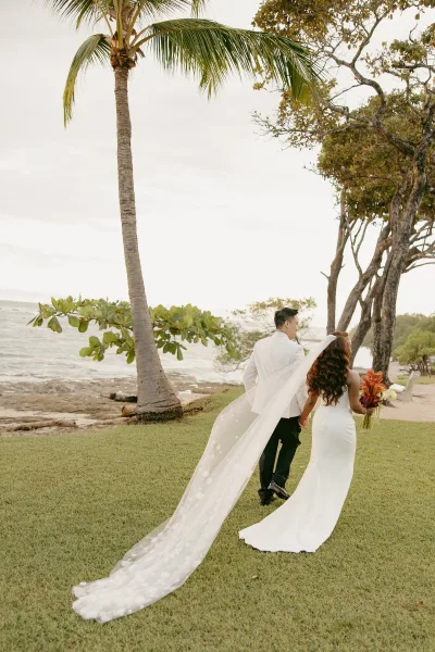 Couple portrait of bride and groom walking away holding hands, her veil train flowing by an ocean shoreline with palm trees under clouds