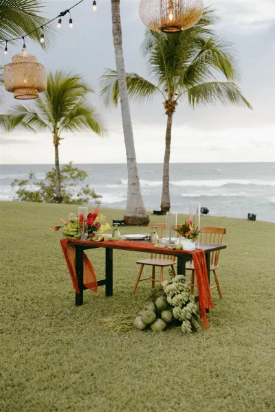 Reception tablescape with tropical wedding tablescape florals, bananas and coconuts on a wood table, under woven lanterns by the ocean and palms