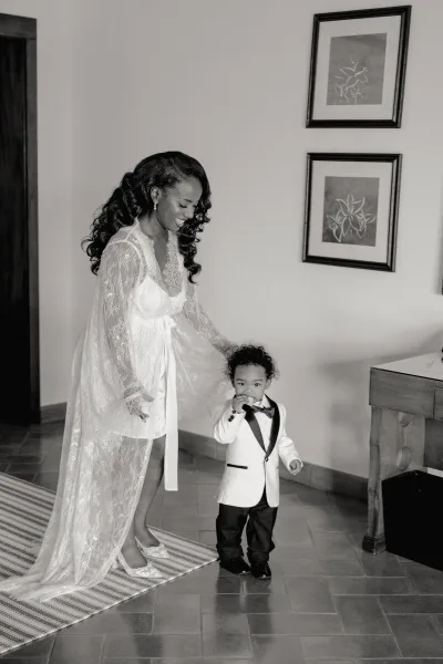 Bridal portrait of bride getting ready in a lace robe with high heels, standing beside a child in tuxedo and bow tie indoors