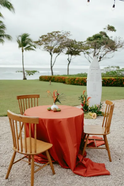Reception tablescape with rust wedding tablecloth on a round table, tropical flowers and fruit, on an oceanfront lawn under string lights