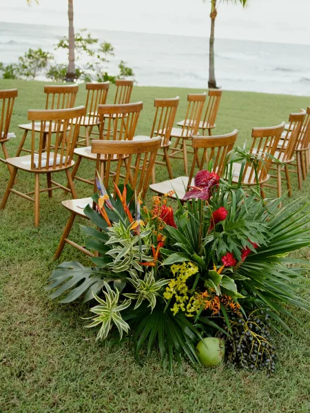 Ceremony setup with outdoor wedding ceremony seating of wood chairs on a lawn, with tropical flowers and palm fronds by the ocean backdrop