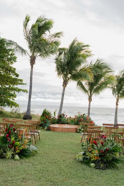 Ceremony setup with beach ceremony setup on a grass lawn, wooden chairs around a wood altar with tropical florals, ocean and palms beyond