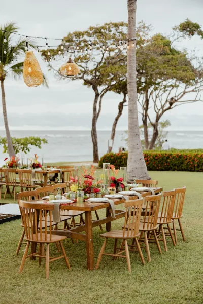 Reception tablescape on a wood farm table wedding setup with tropical floral centerpieces, votive candles, and string lights by the ocean view