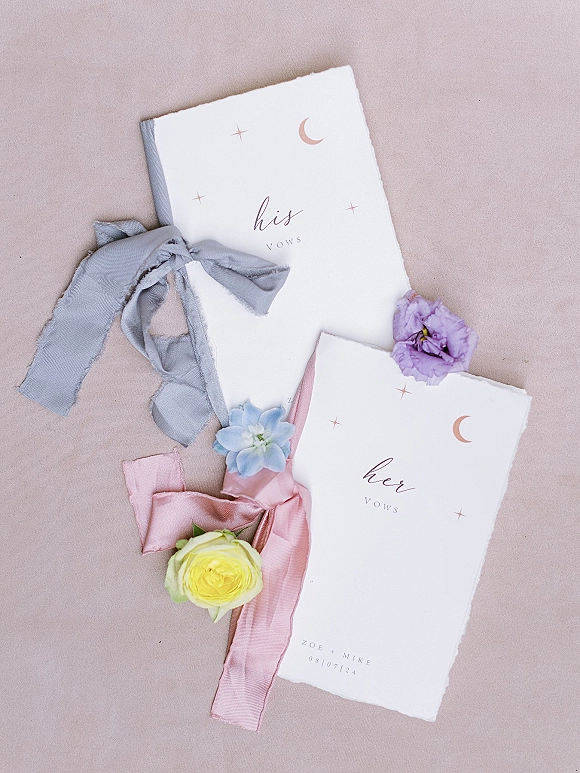 Wedding vows books with his and her vows in torn-edge calligraphy booklets, silk ribbons and small flowers on a neutral fabric backdrop