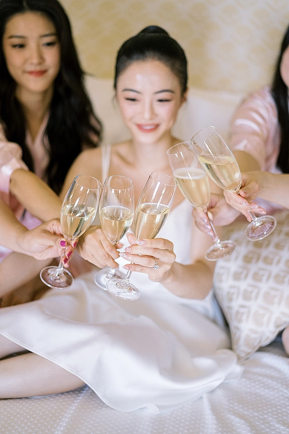 Bridesmaids toast with champagne in pink satin robes, clinking flutes beside the bride’s engagement ring on a bed in the bridal suite