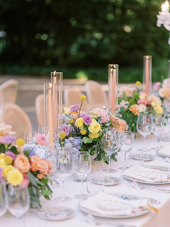 Reception tablescape with pastel wedding centerpiece, taper candles in glass hurricanes, patterned china and glassware on a white linen table outdoors under a chandelier