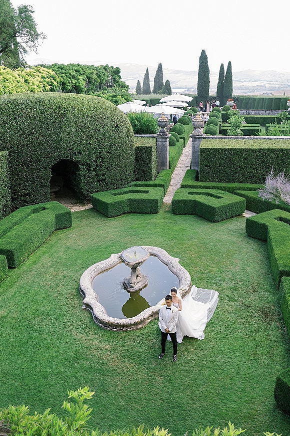Couple portrait of bride behind groom, her veil and long train spread on a stone walkway beside a garden fountain with manicured hedges