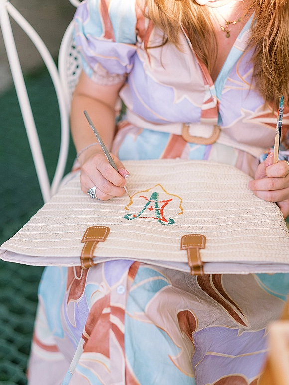 Wedding guest book idea featuring a hand-painted monogram cover beside a straw clutch, paintbrush, and ring near greenery and a chair