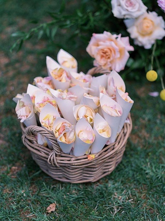 Petal toss cones filled with rose petals in a wicker basket, arranged with roses and greenery on a grass lawn for exit toss