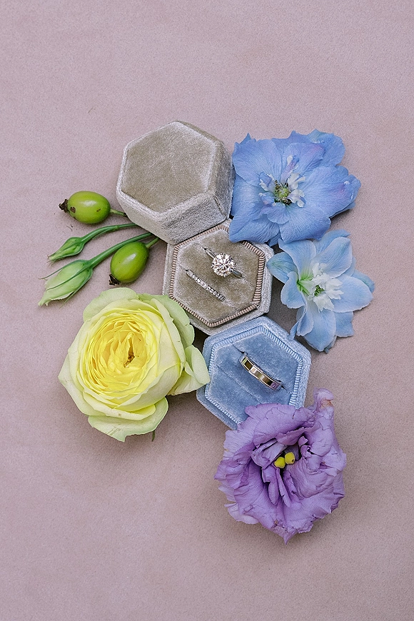 Wedding rings with a round diamond solitaire and pavé band nestled in a velvet ring box beside roses and delphinium on taupe background