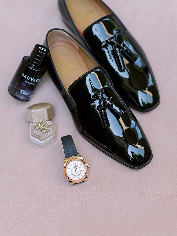 Groom accessories flat lay with black patent loafers, cologne, velvet ring box, gold bands, and watch on blush fabric backdrop