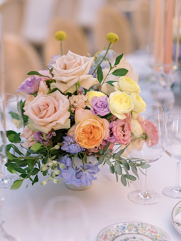 Wedding centerpiece with pastel floral centerpiece of mixed roses and greenery in a low vase on a white tablecloth with wine glasses and floral plate