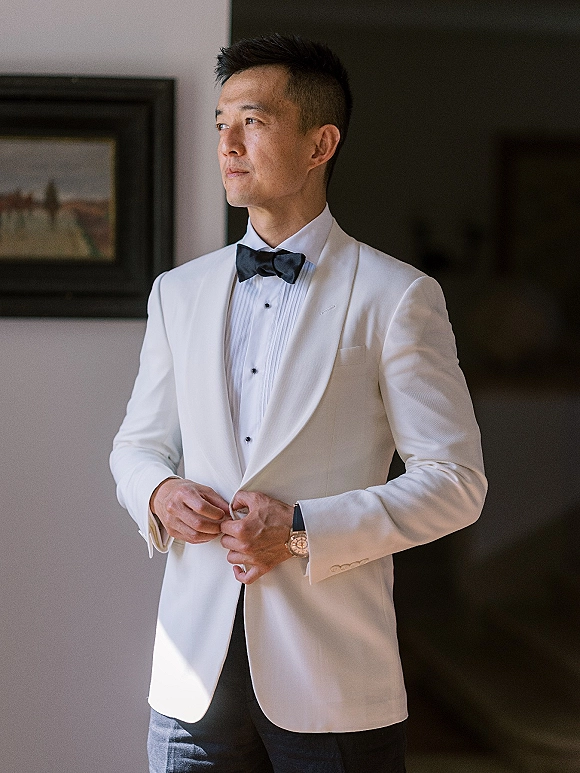 Groom portrait in a white tuxedo jacket and black bow tie, adjusting his jacket with cufflinks and wristwatch indoors by framed art