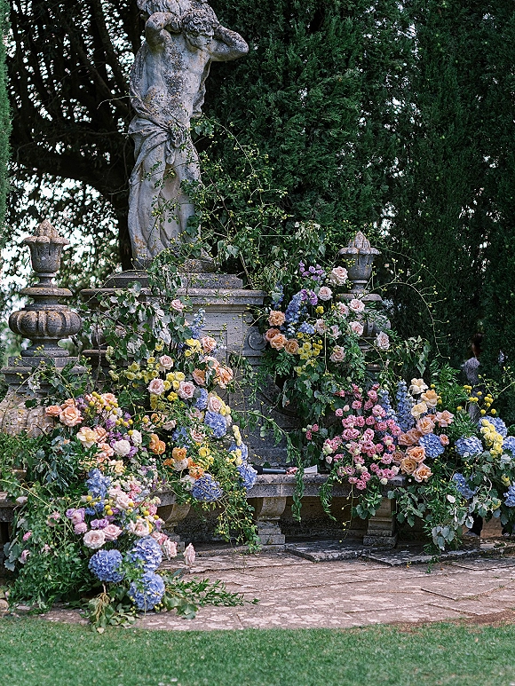 Ceremony altar flowers with lush roses and hydrangeas, climbing greenery, and urn planters on stone steps beside a statue in a garden setting