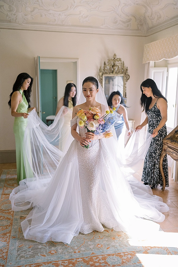 Bride portrait with bridesmaids fixing her bridal veil as she holds a bouquet, standing in window light beside an ornate mirror in a getting ready room