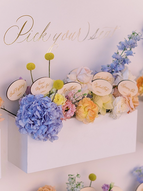 Escort card display with wedding escort cards on a white shelf, topped by a gold calligraphy sign and pastel florals against a white wall