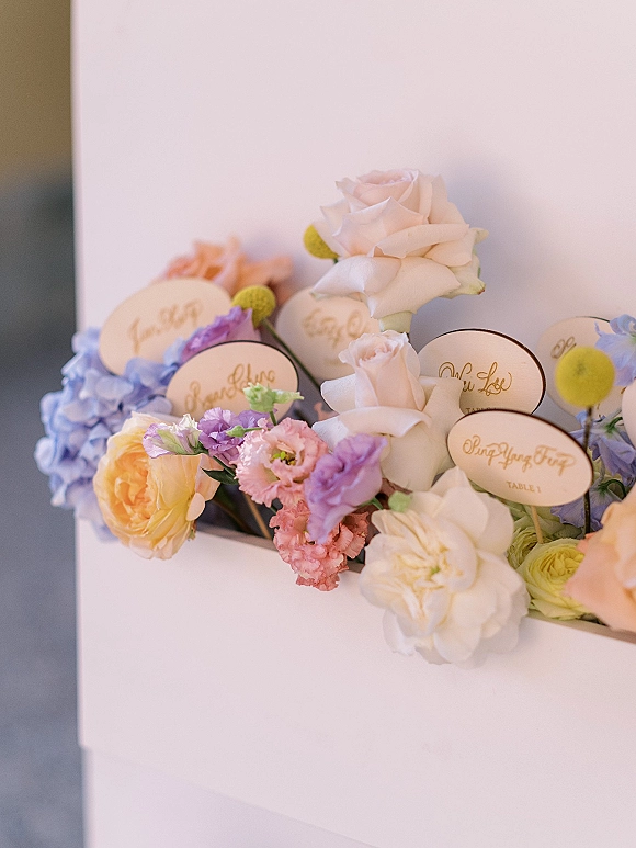 Wedding escort cards with calligraphy on oval place cards nestled in a flower box of roses, hydrangea, and ranunculus on a tabletop
