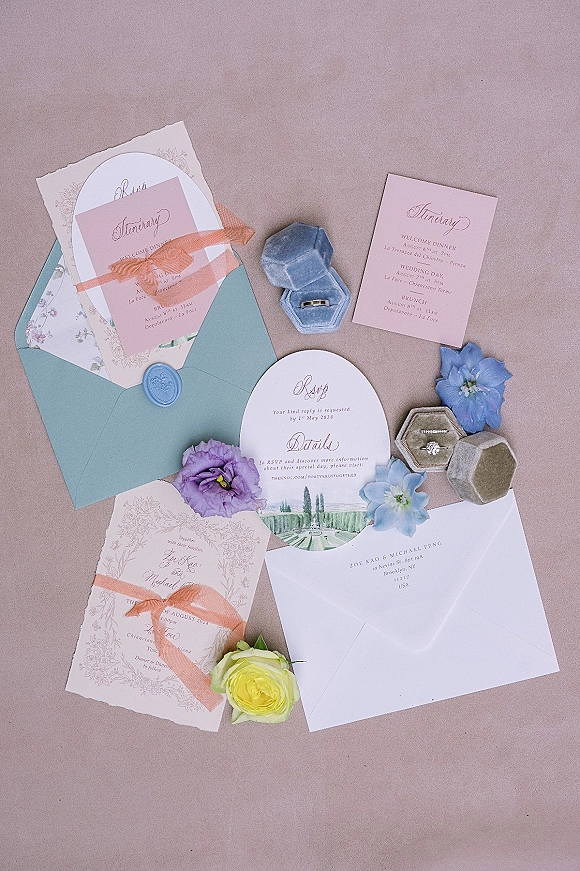 Wedding invitation suite with pastel wedding invitations, blue wax seal and silk ribbon beside rings and florals on a neutral surface