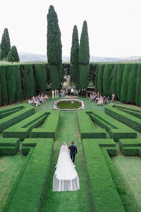 Wedding processional with bride walking down aisle in a wedding dress and cathedral veil as groom escorts her past pond in a formal garden