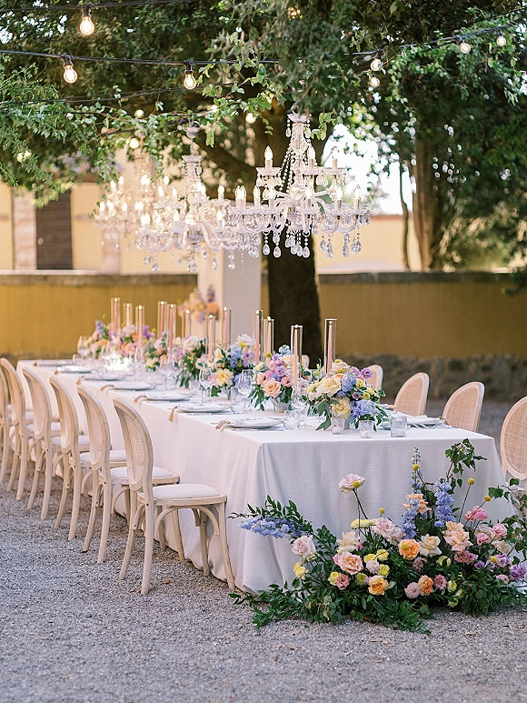 Reception tablescape with outdoor reception table on a long white-clothed banquet, taper candles, florals, chandeliers under trees and string lights