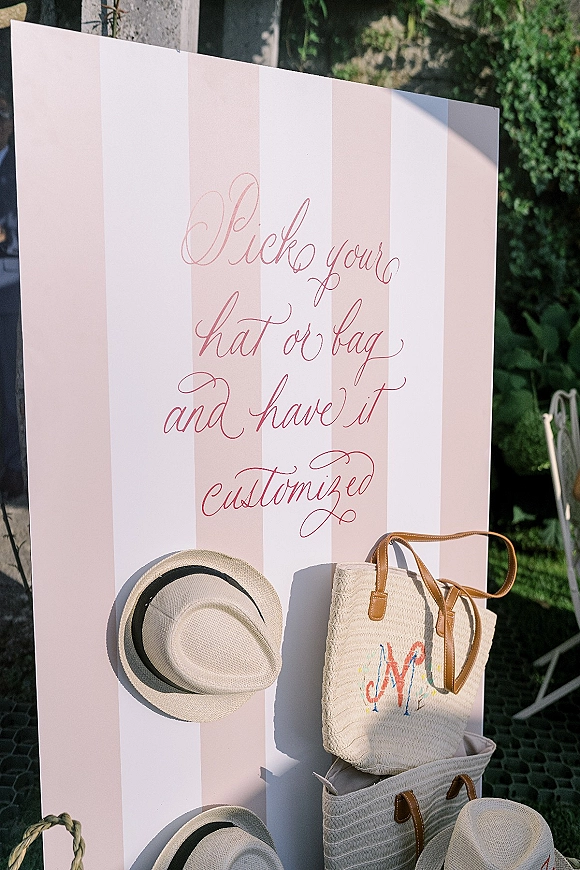 Wedding welcome sign with pick a hat sign calligraphy on a striped board beside straw hats and woven totes on an outdoor patio by greenery