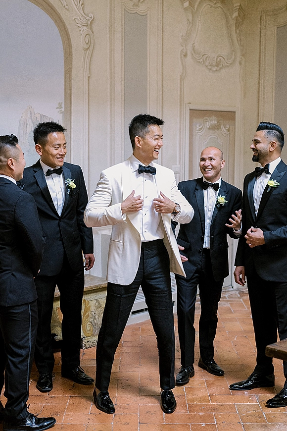 Groomsmen portrait of the groom with groomsmen in black tuxedos and one white dinner jacket, laughing by an ornate arched doorway
