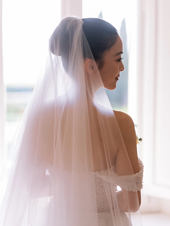 Bridal portrait of a bride in side profile with a sheer veil over her face, standing by a bright window in an indoor room