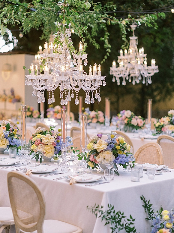 Reception tablescape at an outdoor wedding reception with crystal chandeliers, pastel florals, taper candles, white linens, and string lights among trees