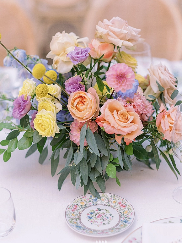 Wedding centerpiece with pastel wedding centerpiece roses, dahlias and eucalyptus on a white tablecloth with floral china and wine glasses on an outdoor patio