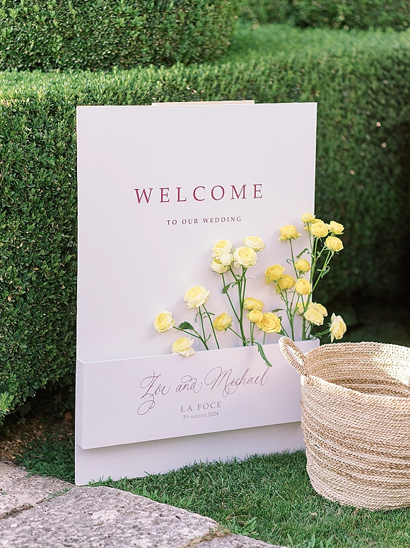 Wedding welcome sign with modern wedding signage calligraphy on a white board, accented by yellow roses in a woven basket by a boxwood hedge