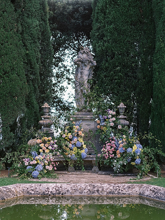 Ceremony altar decor with garden wedding altar stone statue on a pedestal, framed by lush roses, hydrangeas and greenery beside a reflecting pool
