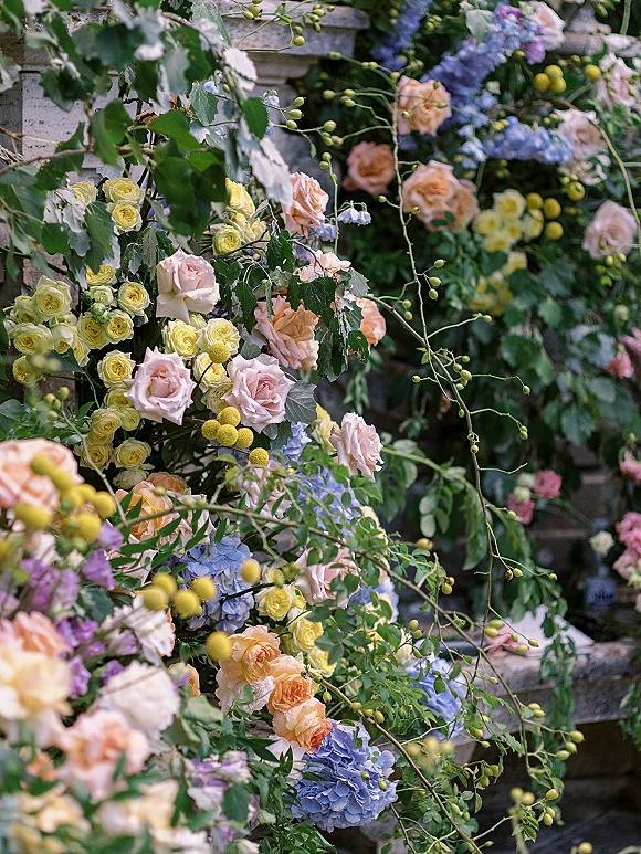 Wedding floral installation and ceremony flower arch with peach roses and blue hydrangea cascading over a stone wall backdrop