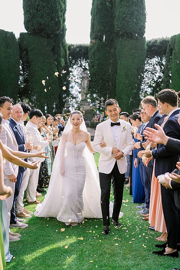 Wedding recessional as bride and groom walking aisle in tuxedo and strapless gown with veil while guests toss rose petals on a garden lawn